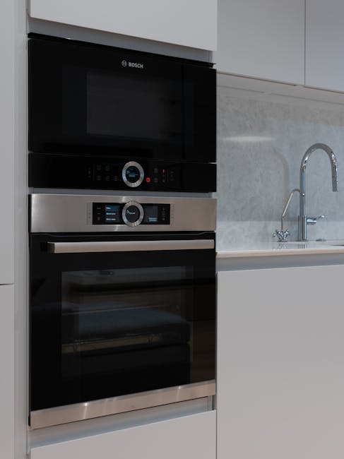 A modern kitchen with a black induction cooktop on a sleek black countertop, clean and shiny white ceramic subway tile splashback, white cabinetry, and built-in appliances including a microwave and ov